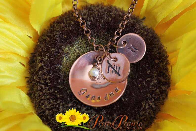 May include: Copper necklace with three charms. The largest charm is round and has the words "Grandma" engraved on it. The middle charm is round and has the Roman numeral "XII" engraved on it. The smallest charm is round and has the number "II" engraved on it. A small pearl bead is attached to the charm with the words "Grandma".