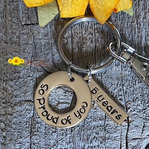 May include: A gold-toned keychain with two charms. One charm is a round disc with the text "So Proud of You" and the other charm is a rectangular tag with the text "5 Years".
