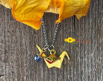 May include: Gold origami crane necklace with a blue crystal charm. The necklace is on a silver chain and is hanging from a wooden surface.