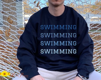 Op de afbeelding: Een zwart sweatshirt met het woord "SWIMMING" vier keer herhaald in een blauwe gradiënt.