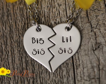 May include: Two silver necklaces with heart-shaped pendants that say "Big Sis" and "Lil Sis". The pendants are broken in half, symbolizing a sisterly bond.