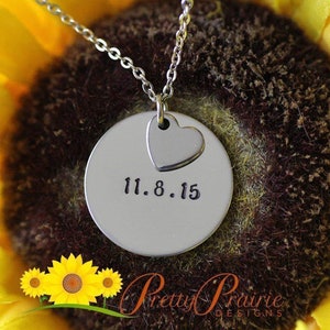 May include: A silver necklace with a round pendant featuring the date "11.8.15" and a small heart charm. The necklace is photographed against a yellow sunflower.