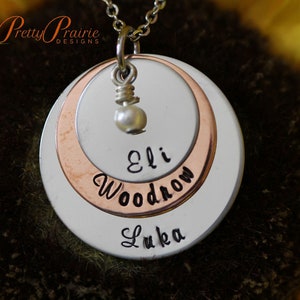 May include: A silver and copper layered necklace with a pearl charm. The necklace has three names engraved on it: Eli, Woodrow, and Luka.