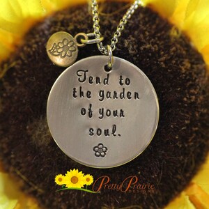 May include: Silver necklace with a round pendant that says "Tend to the garden of your soul." The pendant is hanging from a silver chain and has a small flower charm attached to it. The necklace is photographed against a yellow sunflower.