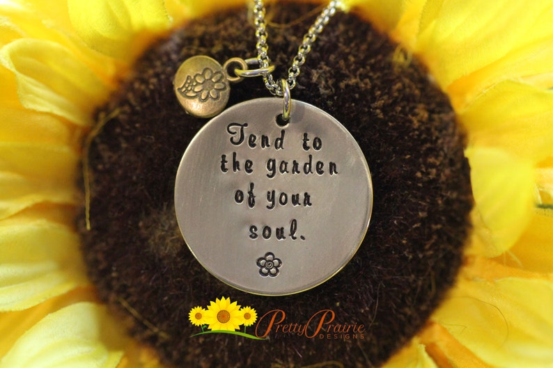 May include: Silver necklace with a round pendant that says "Tend to the garden of your soul." The pendant is hanging from a silver chain and has a small flower charm attached to it. The necklace is photographed against a yellow sunflower.