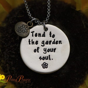 May include: Silver necklace with a round pendant that says "Tend to the garden of your soul." The pendant has a small flower design on it. The necklace is on a dark background.