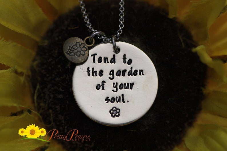 May include: Silver necklace with a round pendant that says "Tend to the garden of your soul." The pendant has a small flower design on it. The necklace is on a dark background.