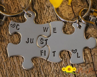 May include: Two silver puzzle pieces with key rings attached. The pieces fit together and have the words "We Just Fit" engraved on them. Each piece also has a small heart symbol.