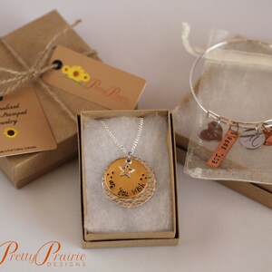May include: Two gift boxes with jewelry inside. One box contains a silver necklace with a gold charm that says "as you wish" and a star. The other box contains a silver bangle bracelet with charms that say "Est. 1998" and "M" and two heart charms.