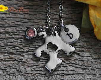 May include: A silver-toned necklace featuring a puzzle piece charm with a heart-shaped cutout, a small round charm stamped with the letter D, and a circular pink crystal charm, all suspended from a delicate silver-toned metal chain.