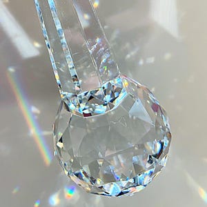May include: A clear, faceted crystal pendant featuring a spherical base attached to a long, tapered, multi-faceted stem. The crystal refracts light, creating rainbow-colored reflections. A small heart-shaped tag with the letters E&M&D is attached.