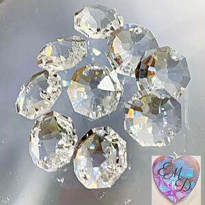 May include: A collection of clear, faceted crystal beads with an iridescent sheen, scattered on a reflective surface. The beads are octagonal and reflect light, creating a rainbow effect. A heart-shaped charm with the letters "EMD" is in the bottom right corner.