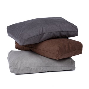 May include: Three rectangular dog beds stacked on top of each other. The top bed is dark gray, the middle bed is brown, and the bottom bed is light gray.