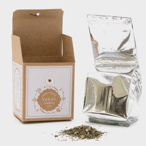 May include: A brown cardboard box with a white label that says "Sarah's Garden Tea" and a silver foil pouch of tea. The tea is loose leaf and is scattered on a white surface.
