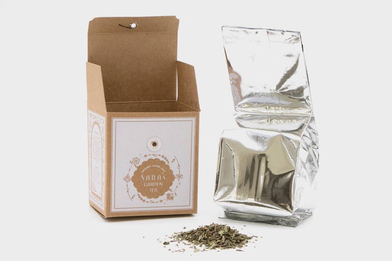 May include: A brown cardboard box with a white label that says "Sarah's Garden Tea" and a silver foil pouch of tea. The tea is loose leaf and is scattered on a white surface.
