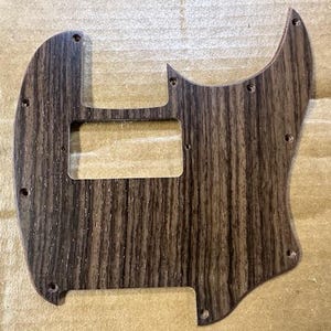 May include: A guitar pickguard with a dark wood grain pattern. The pickguard has a rectangular cutout for the pickups and several screw holes. The wood grain has a vertical pattern with dark and light brown streaks.
