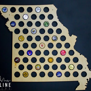 May include: A wooden cutout of the state of Missouri with 100 circular holes for displaying bottle caps. The holes are filled with various colored bottle caps, including red, blue, green, yellow, and silver. The board is made of light brown wood and has a natural wood finish.