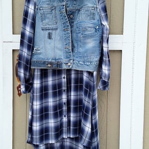 May include: A denim vest with distressed details is layered over a long-sleeved, button-down shirt. The shirt and vest are hanging on a white hanger. The shirt features a blue and white plaid pattern. The vest has multiple pockets.