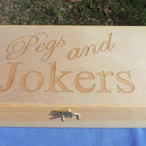 Peut inclure: Une boîte en bois de couleur claire avec les mots "Pegs and Jokers" gravés sur le couvercle. La boîte a un fermoir en métal et est posée sur une surface bleue. Un jeu de cartes est visible.