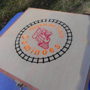 May include: A wooden dominoes game box with a light wood finish. The lid features a circular design with the words "Mexican Train Dominoes" in orange, surrounding a red train illustration. The design is framed by a black railroad track.