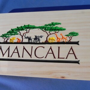 May include: Wooden Mancala game board with a carved African savanna scene. The design features green trees, orange giraffes, yellow and orange animals, and gray elephants above the word "MANCALA" in brown.