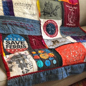 May include: A patchwork quilt made from repurposed t-shirts. The quilt features a variety of colors and designs, including a blue and white "Save Ferris" t-shirt, a red and white "Rocky Mountain" t-shirt, and a green and white "Centaurus Theatre Company" t-shirt. The quilt is stitched together with a white thread.