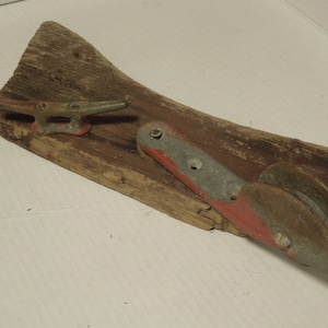 May include: A weathered piece of driftwood with two metal boat cleats attached. One cleat is a simple hook, the other is a pulley with a red and gray paint job.