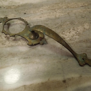 May include: An antique brass door handle with an ornate design. The handle has a curved grip and a decorative backplate with a circular opening. The brass has a weathered, aged appearance, with hints of green oxidation.