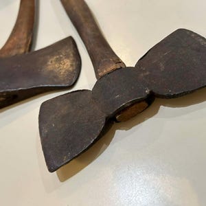 May include: Two vintage axes with wooden handles and dark, weathered metal blades. One axe has a single blade, while the other has a double-sided blade. The axes are likely used for chopping wood or other similar tasks.