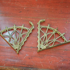 May include: Two ornate gold metal shelf brackets with a decorative fan-shaped design. The brackets are made of cast iron and have a distressed finish.