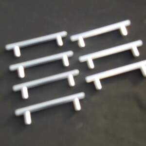May include: A collection of seven silver-colored cabinet pulls with a T-shaped design. The pulls are arranged on a dark surface, showcasing their simple, modern aesthetic. These hardware pieces are suitable for drawers and cabinets.
