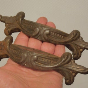 May include: Two antique cast iron drawer pulls with ornate scrollwork designs. The pulls are a dark brown color and have a distressed finish.