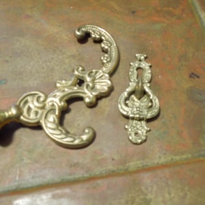 May include: Two ornate brass hooks with intricate designs. One hook is a single hook with a decorative base. The other hook is a double hook with a decorative loop.
