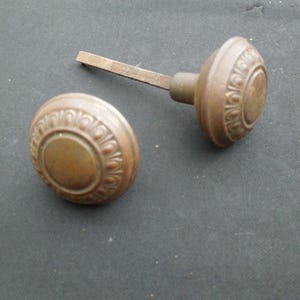 May include: Two antique bronze door knobs. One knob is attached to a metal rod, while the other is separate. Both knobs have a circular design with a central circle and an ornate border. The knobs are a warm, aged bronze color.