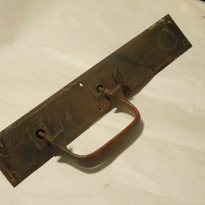 May include: Antique brass door handle with a red metal pull handle. The handle has a rectangular plate with two keyholes and a circular design.