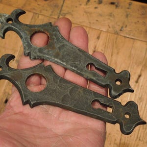 May include: Two antique metal door or cabinet hardware pieces with ornate fleur-de-lis designs and keyhole cutouts. The pieces are a dark gray color with a rustic finish.