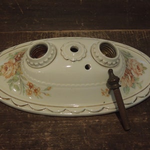 May include: A vintage porcelain ceiling light fixture with three sockets and a floral design. The fixture is oval shaped and has a gold trim. It is in good condition and would be a great addition to any vintage home.