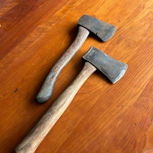 May include: Two hand axes with weathered metal heads and wooden handles. The top axe features a curved handle and a dark, oxidized steel blade. The bottom axe has a straight wooden handle and a slightly wider, worn metal head with visible surface texture.