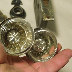 May include: Two clear glass door knobs with a starburst pattern in the center. The knobs are held in a hand.