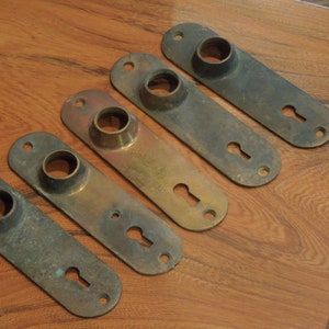 May include: Five antique brass door plates with keyholes and a round hole for a knob. The plates are oval shaped and have a worn, aged patina.