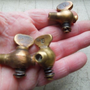 May include: A collection of vintage brass spigots with a screw-in design. Each spigot has a small, oval handle with a letter stamped on it. The spigots are a warm, antique gold color and have a spring mechanism.
