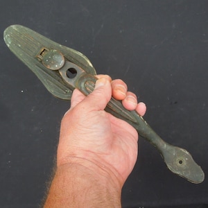 May include: A green metal door handle with a decorative design. The handle has a small hole in the center and a latch on the side.