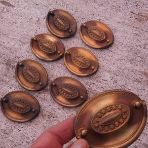 May include: Eight antique oval-shaped brass drawer pulls with ornate designs. The pulls have a slightly aged, tarnished finish.