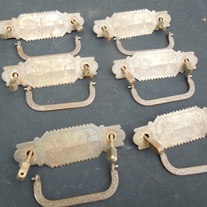 May include: Six antique brass drawer pulls. Each pull features a rectangular backplate with a decorative, saw-tooth edge and a curved handle. The backplates have an etched design of a ship and palm trees. The pulls are tarnished with age.