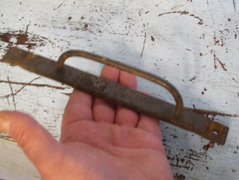 Może przedstawiać: Metalowy uchwyt zardzewiały z pętlą do drzwi lub szuflady. Uchwyt ma około 38 cm długości i ma szorstką, fakturowaną powierzchnię.