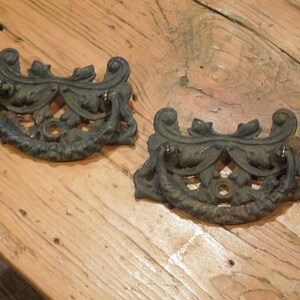 May include: Two antique, ornate, black metal drawer pulls with a scrolled design. The pulls are on a wooden surface.
