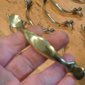 May include: A set of six gold-toned cabinet door pulls with a simple, elegant design. The pulls are made of metal and have a smooth, polished finish.