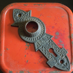 May include: A black metal door knob backplate with intricate floral designs and a keyhole. The backplate is on a red surface.