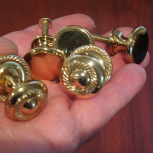 May include: Four gold-toned cabinet knobs with a rope design. Two of the knobs have a round shape, and two have a more intricate design with a raised center.