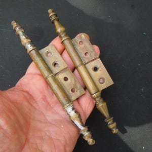 May include: Two antique brass hinges with decorative finials. The hinges are closed and have a worn, aged finish.